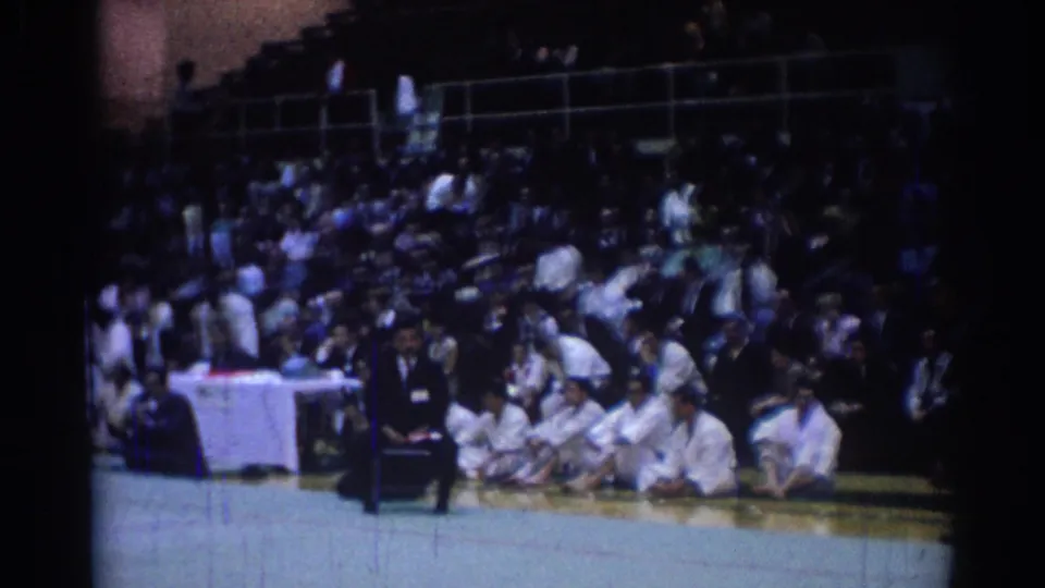 1967 Los Angeles judo competition view showing a dark-suited official beside seated athletes and a large audience in the stands.