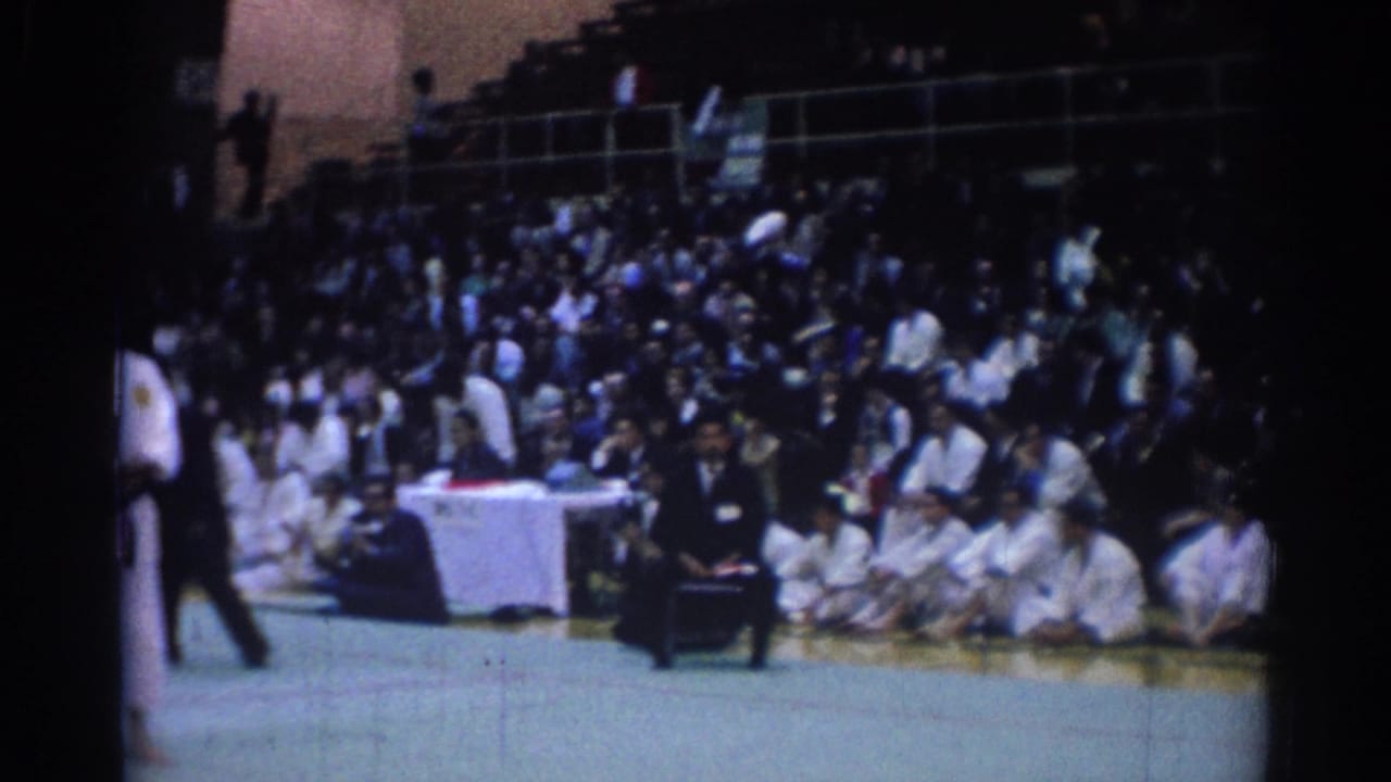 Crowd seated behind a judges' table near a martial arts mat.