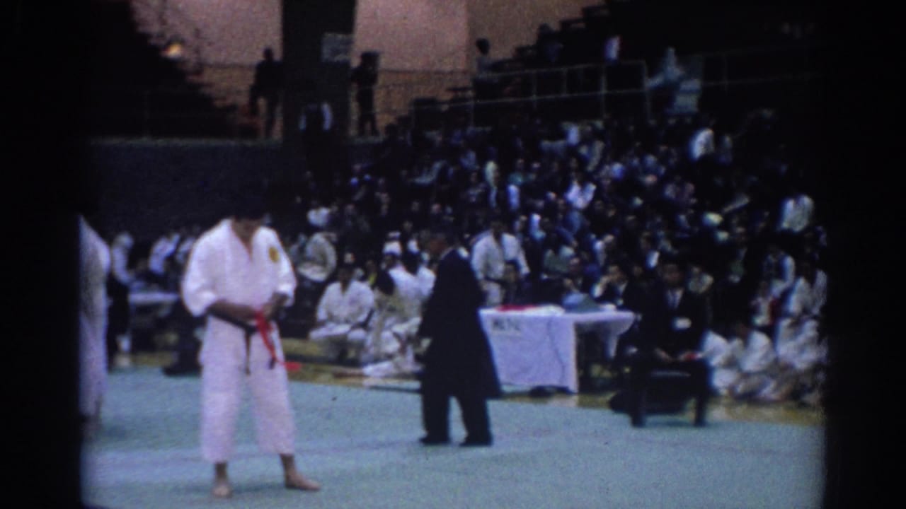 1967 Los Angeles footage showing a martial arts competitor in a white gi with a red belt standing before officials and spectators.