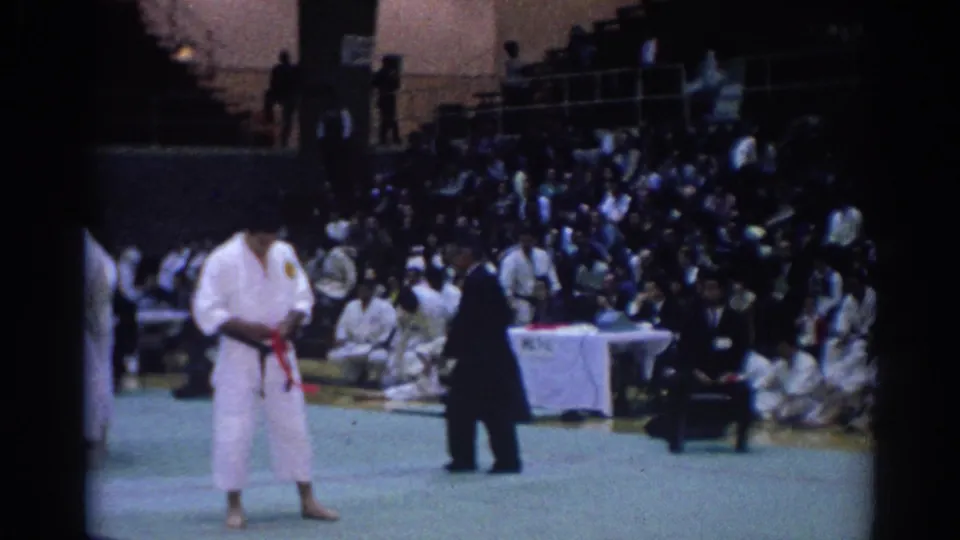 1967 Los Angeles footage showing a martial arts competitor in a white gi with a red belt standing before officials and spectators.