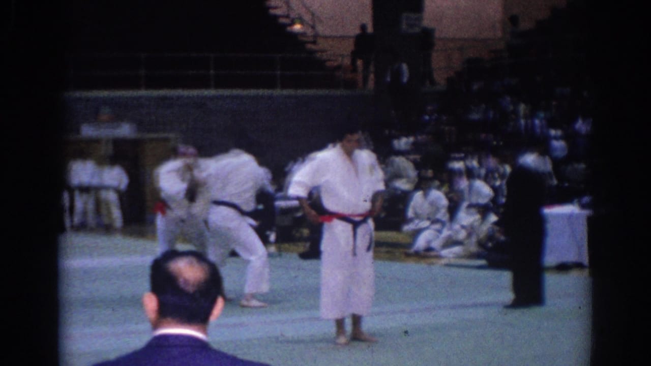 1967 Los Angeles martial arts tournament scene with white-gi competitors, colored belts, a referee, and spectators around the mat.