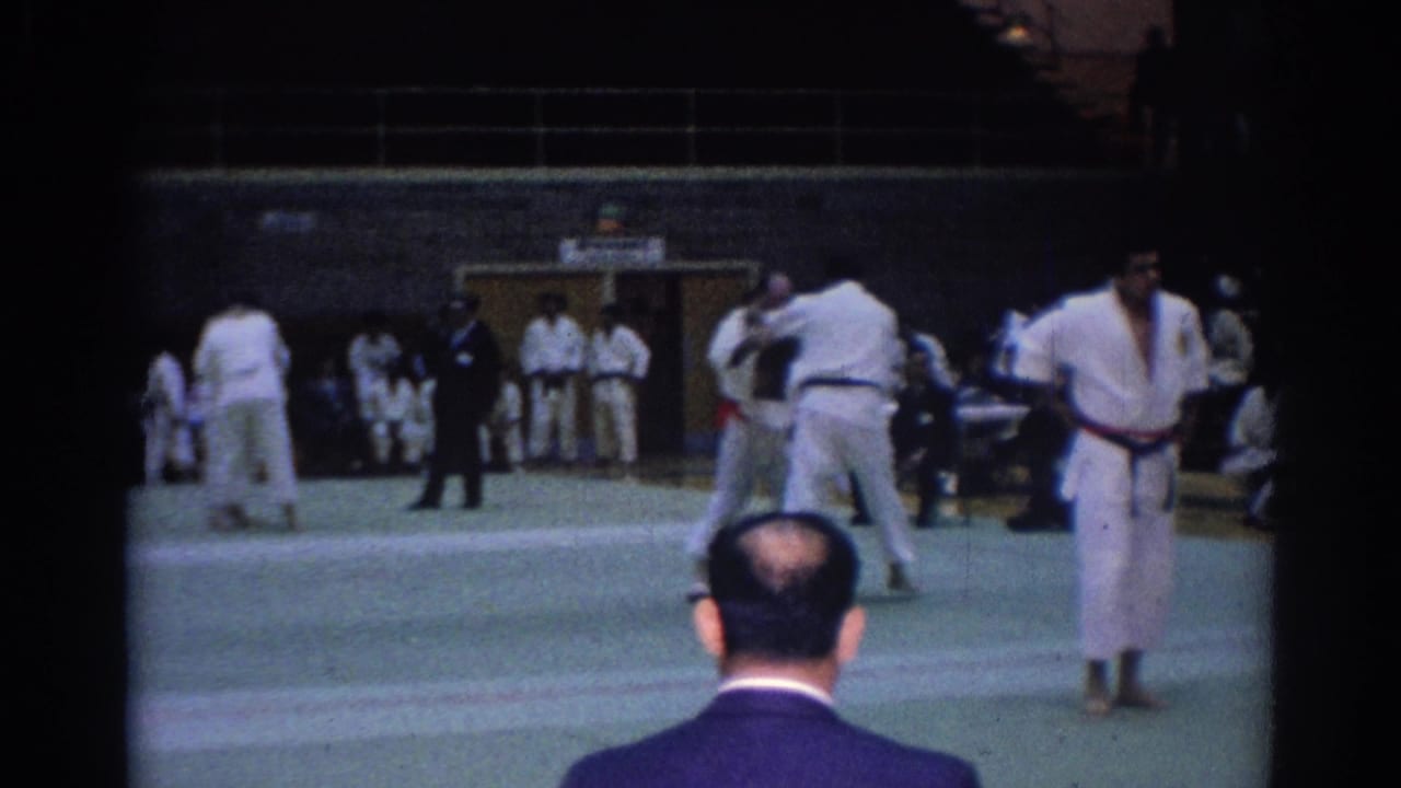 Wide 1967 Los Angeles tournament view with competitors in gi, officials, spectators, and a foreground viewer's head at the mat edge.