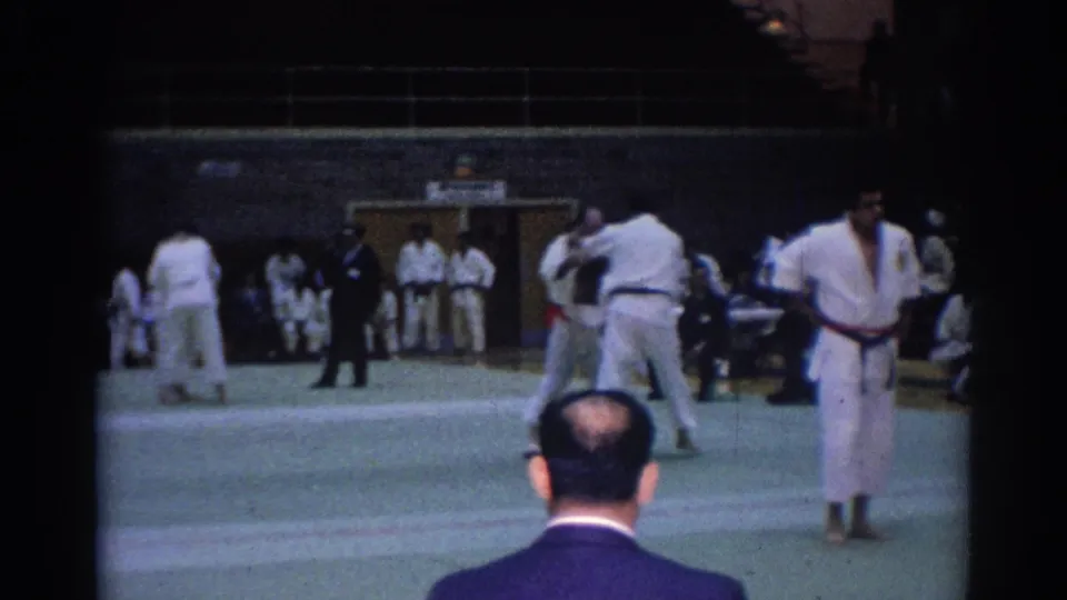 Wide 1967 Los Angeles tournament view with competitors in gi, officials, spectators, and a foreground viewer's head at the mat edge.