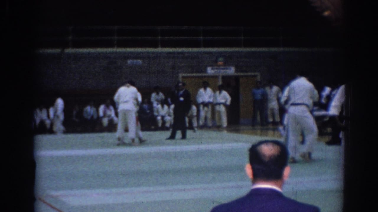 1967 Los Angeles gymnasium footage showing karate competitors on a green mat while a man in a grey suit watches from the foreground.