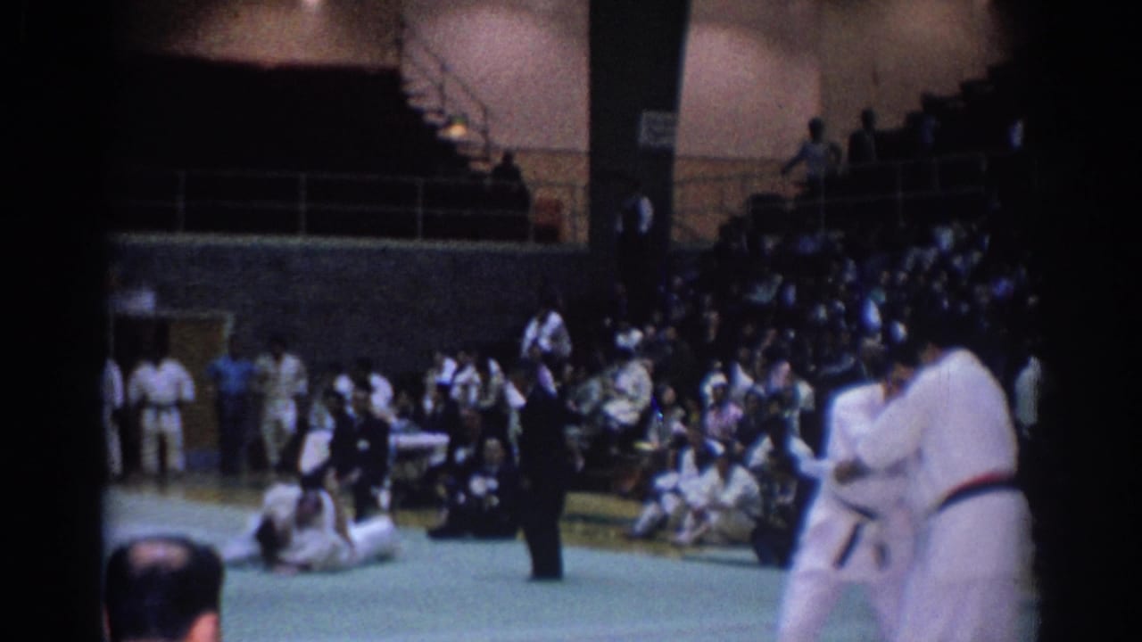 Karate competitors at the edge of the mat with spectators in bleachers.