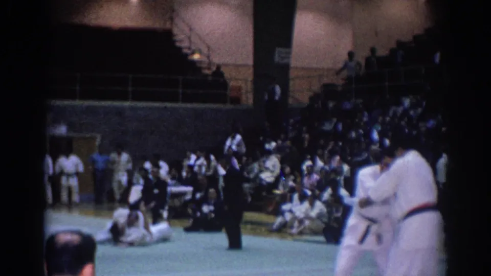 A karate bout in a Los Angeles gymnasium, with a referee standing near the mat and spectators filling the bleachers behind.