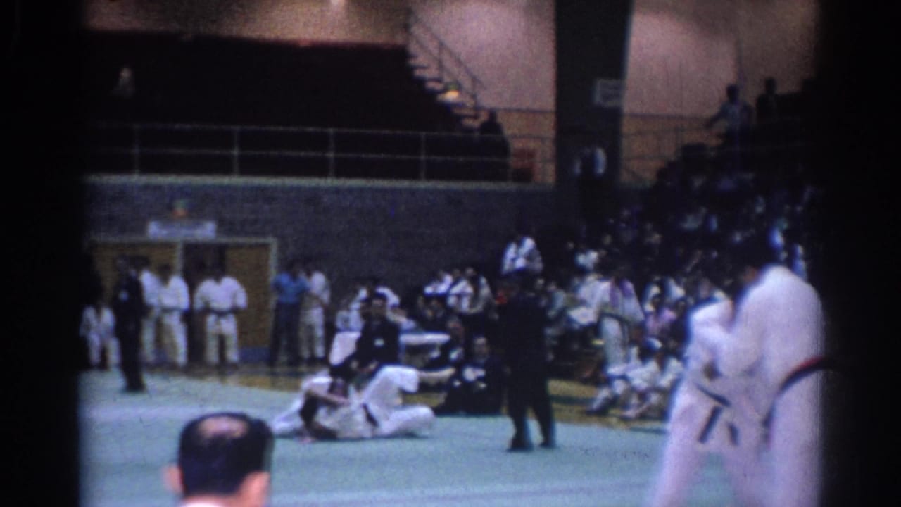 Home-movie view of a karate match with two gi-clad competitors blurred by motion and a seated audience along the gym wall.