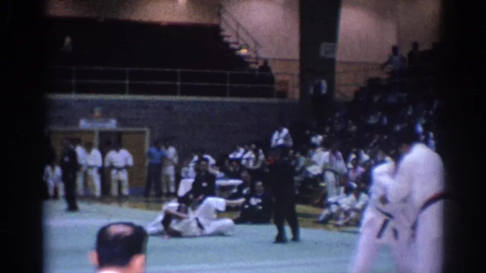 Home-movie view of a karate match with two gi-clad competitors blurred by motion and a seated audience along the gym wall.