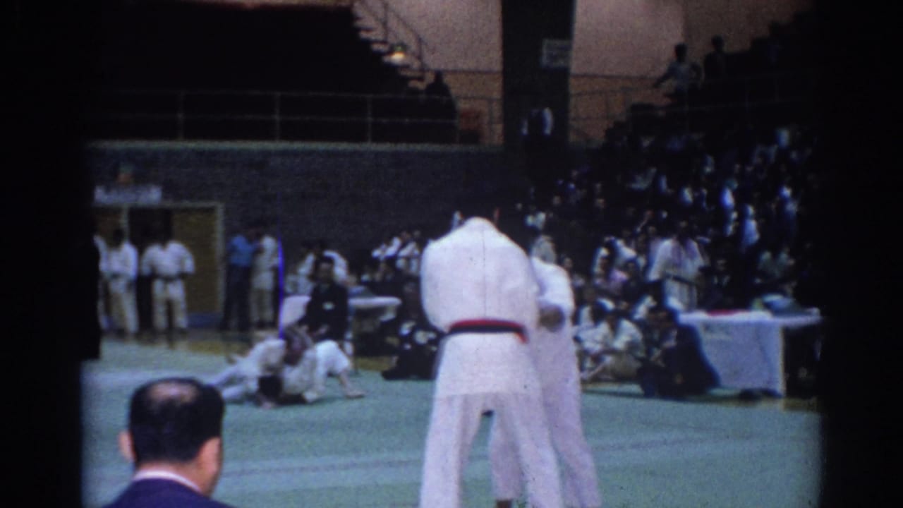 Two karate competitors in white gi face each other on a green mat while seated teams and spectators watch inside the gymnasium.