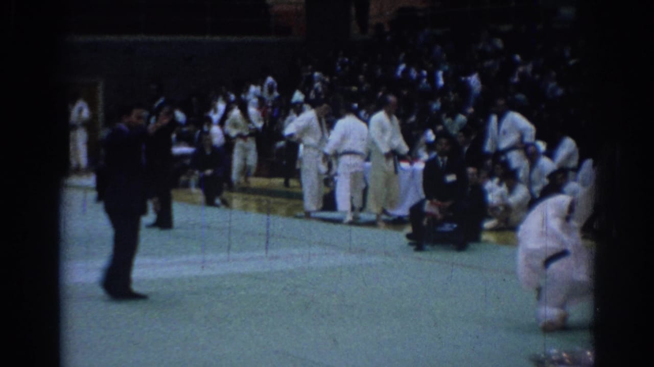 Karate participants gather near a mat as spectators sit in bleachers.