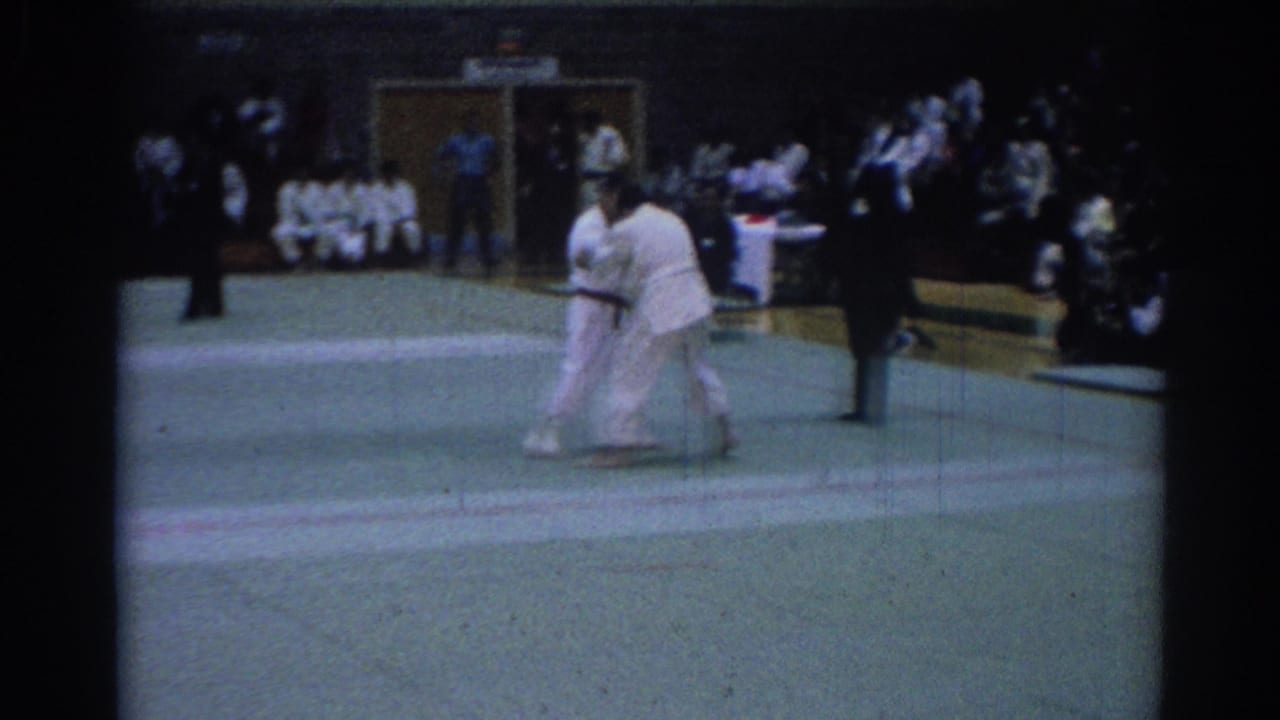 Two competitors in white gi grapple on a green mat during a 1967 Los Angeles indoor karate match, with spectators in the background.