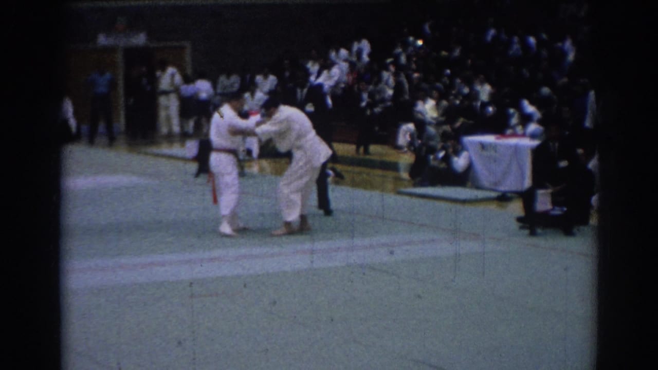 Karate competitors in white gi face off on a gym mat in Los Angeles, with a judge table and seated crowd visible behind them.