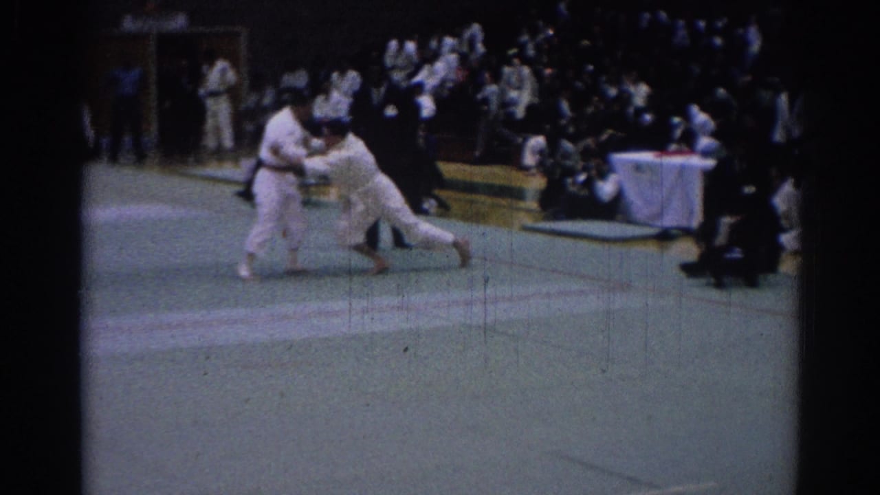 A 1967 indoor karate match moment shows one competitor pushing forward as the other braces, with spectators and officials nearby.