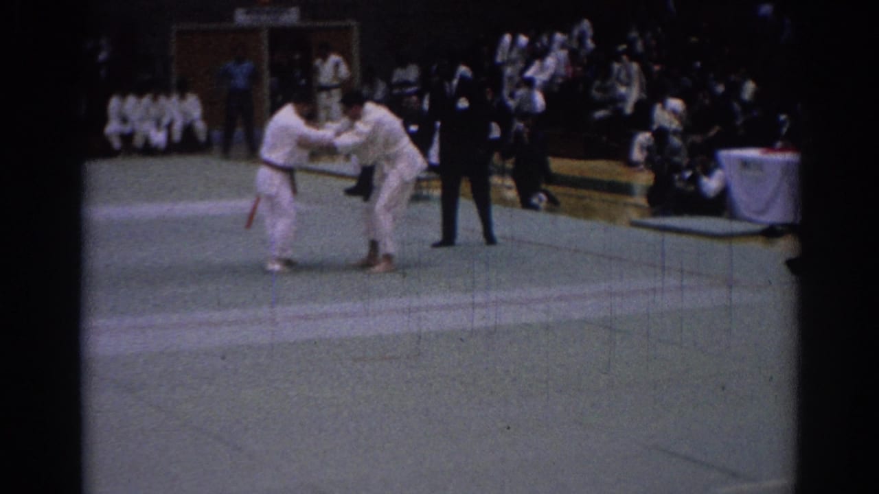 Two martial arts competitors in white gi grapple on a green mat as spectators watch.