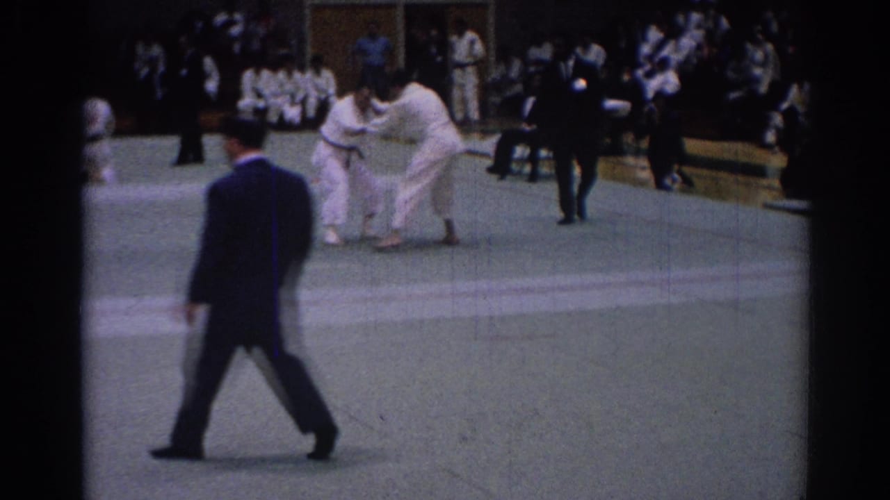 A dark-suited referee walks across the foreground as two white-uniformed competitors grapple in the tournament area.