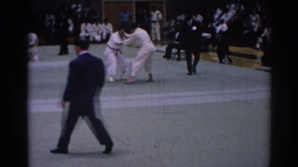 A dark-suited referee walks across the foreground as two white-uniformed competitors grapple in the tournament area.