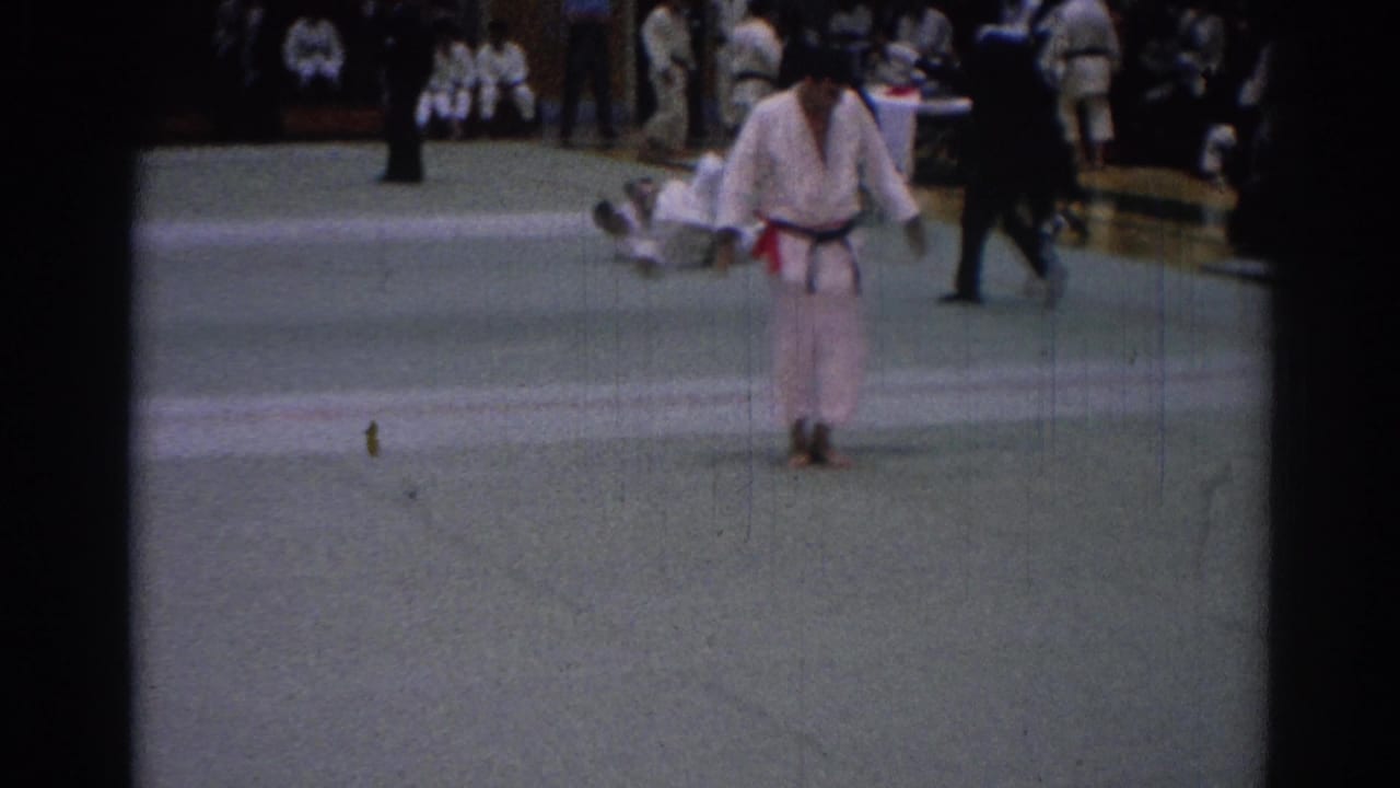 One competitor in a white gi and dark belt walks toward the camera after action on the green mat, with another figure down behind.