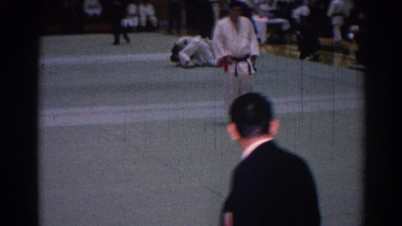 A white-uniformed competitor stands close to the camera, with the match area and a downed participant visible behind him.