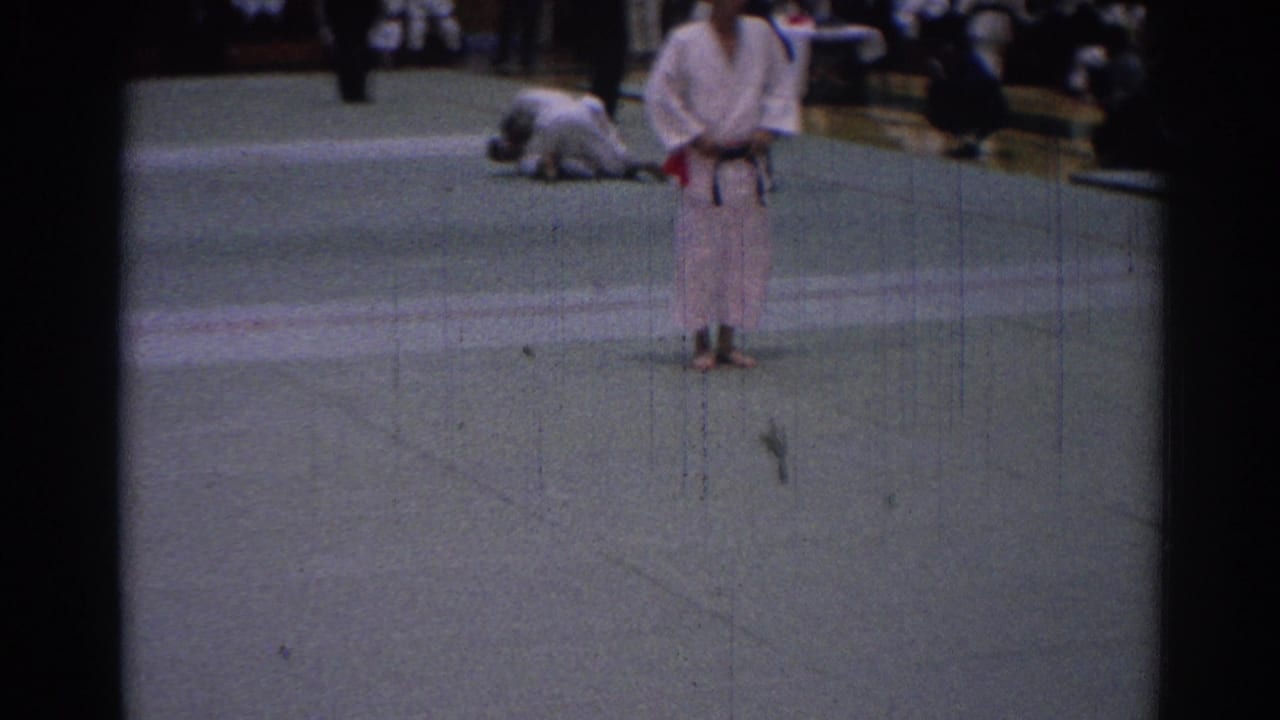 Judo competitor in a white gi stands on a mat while another competitor is down behind him.