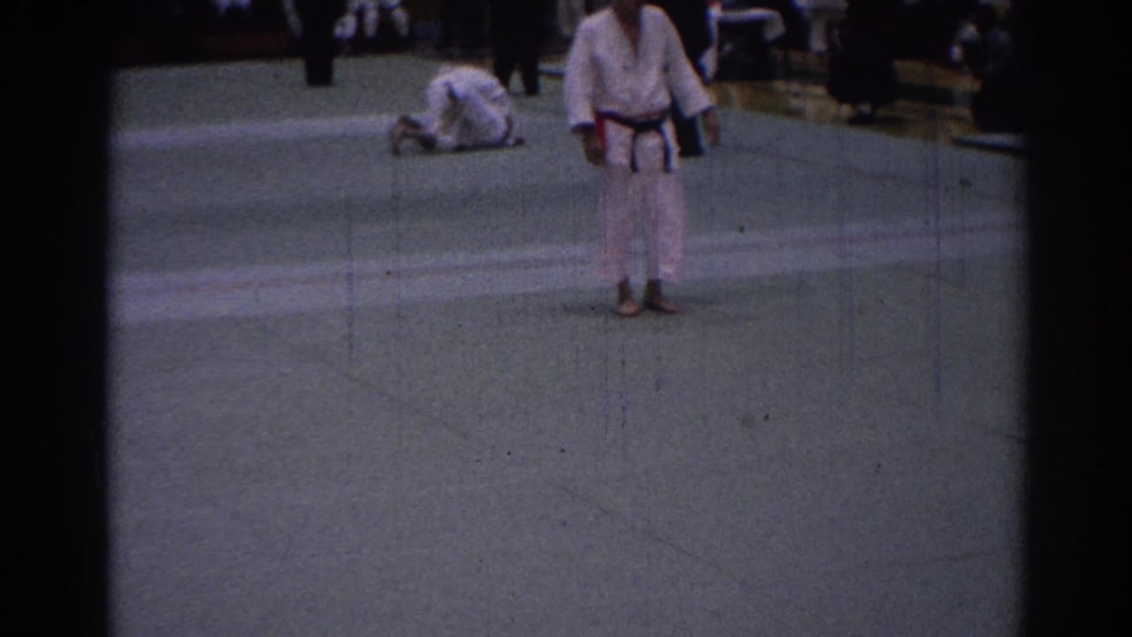 A white-gi judoka stands in an indoor Los Angeles gym as another competitor is on the mat, with spectators visible beyond the action.