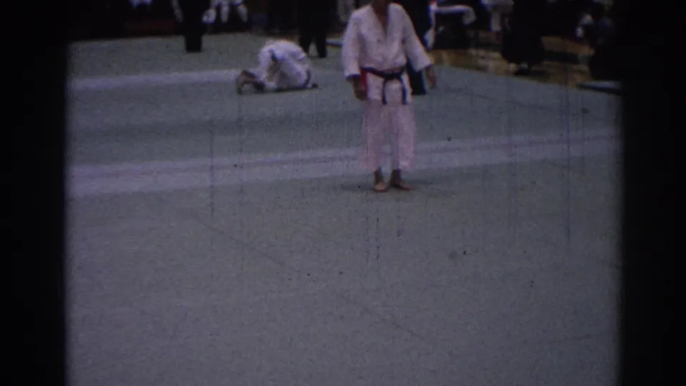 A white-gi judoka stands in an indoor Los Angeles gym as another competitor is on the mat, with spectators visible beyond the action.
