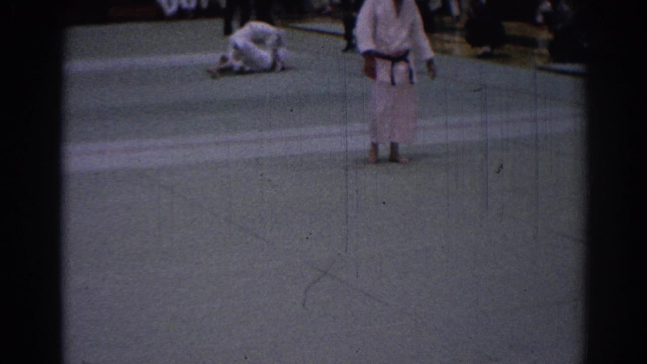 Handheld 8mm footage shows a judo competitor standing near the foreground while a second fighter lies or kneels on the mat in a gym setting.