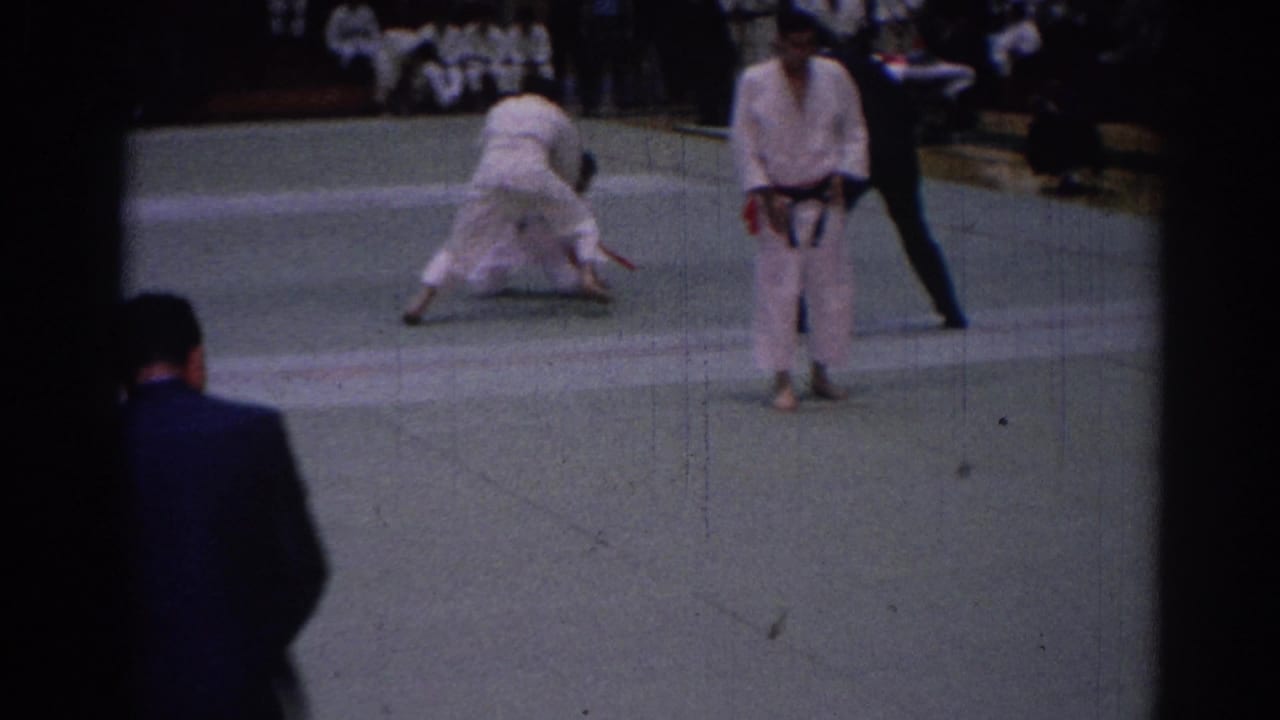 In the clearest action frame, two judoka engage on the mat as a dark-suited referee observes beside a standing competitor and seated spectators.