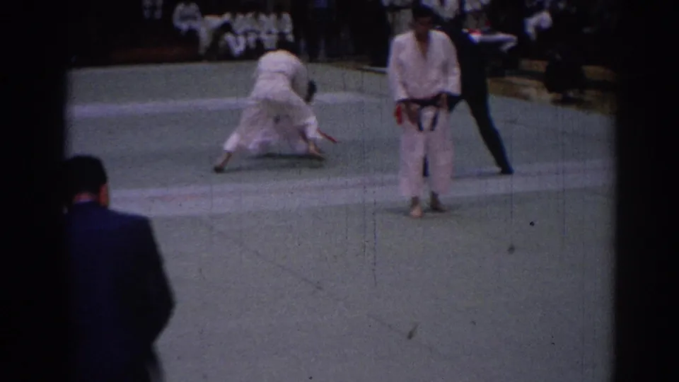 In the clearest action frame, two judoka engage on the mat as a dark-suited referee observes beside a standing competitor and seated spectators.