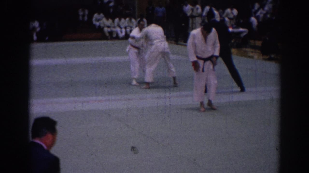 Two competitors in white gi spar on an indoor mat while a referee stands nearby.