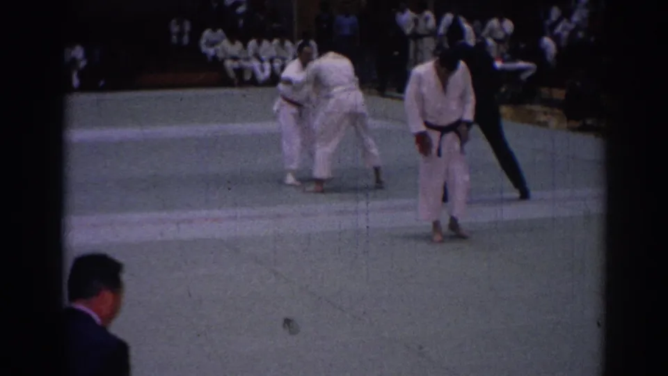 1967 Los Angeles 8mm frame showing two karate competitors on a green mat with a referee and seated spectators in the background.