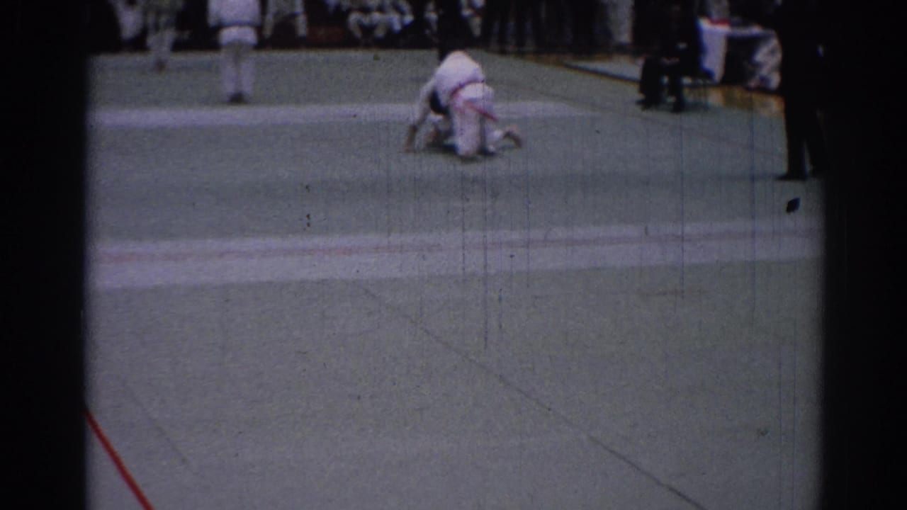 1967 Los Angeles 8mm frame showing a takedown moment on the indoor mat, with uniformed spectators visible behind.