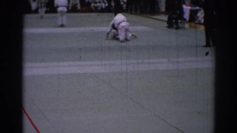 1967 Los Angeles 8mm frame showing a takedown moment on the indoor mat, with uniformed spectators visible behind.