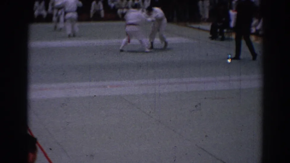 1967 Los Angeles karate tournament frame with two gi-clad competitors grappling and a referee moving along the right edge.