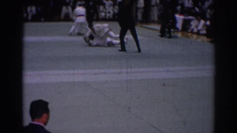 1967 Los Angeles 8mm frame showing karate competitors in white gi uniforms on a green mat as an official watches.
