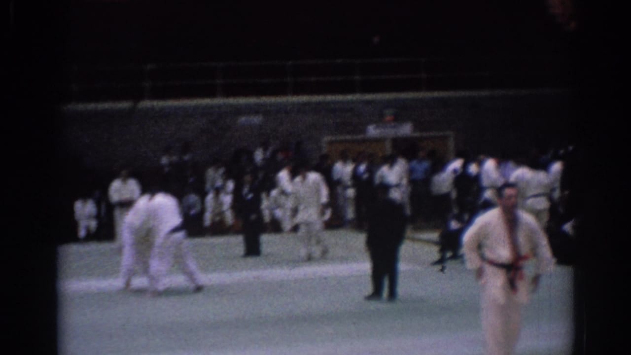 Wide 8mm frame from a 1967 Los Angeles karate competition with competitors in gi uniforms, officials, and a crowd at the edge of the mat.