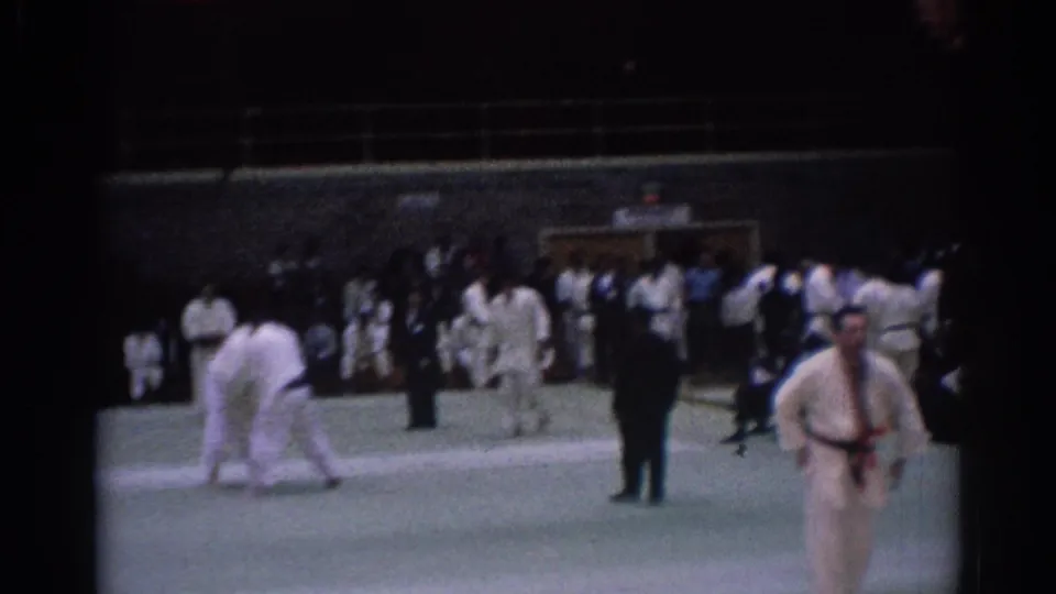 Wide 8mm frame from a 1967 Los Angeles karate competition with competitors in gi uniforms, officials, and a crowd at the edge of the mat.