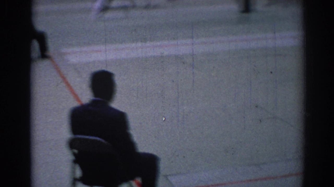 1967 Los Angeles 8mm frame with a seated foreground figure watching blurred karate action across the green mat.
