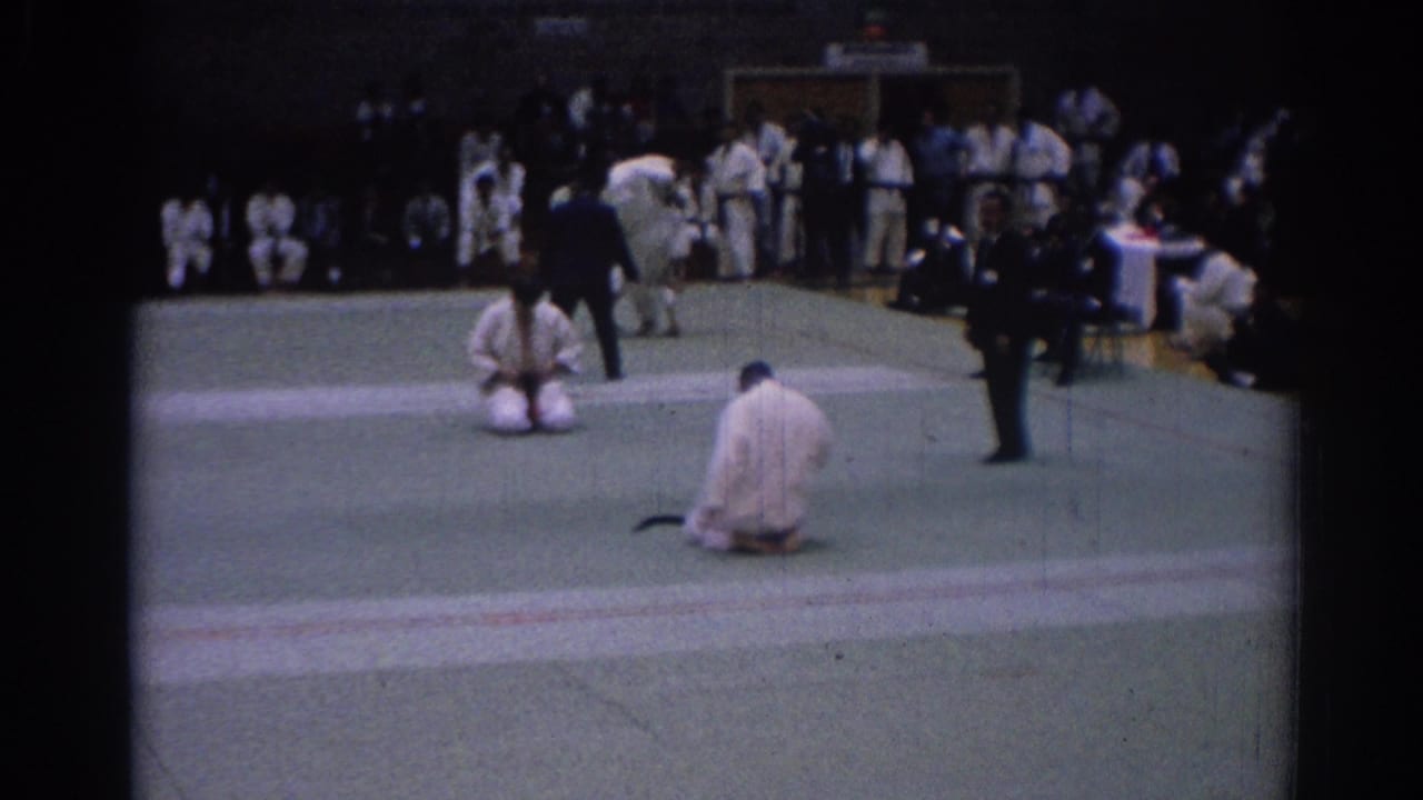 Two karate competitors kneel on separate mat areas while spectators stand along a gym wall.