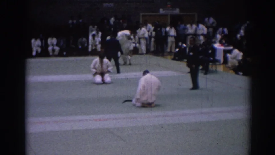 1967 Los Angeles 8mm frame showing indoor karate competitors kneeling on green mats with officials and spectators nearby.