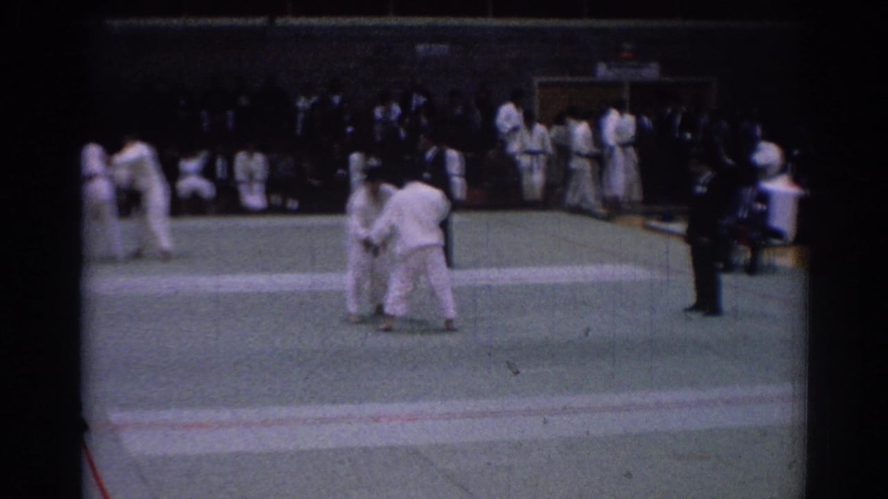 Indoor 1967 Los Angeles karate match frame with competitors in motion, a nearby official, and spectators along the gym edge.