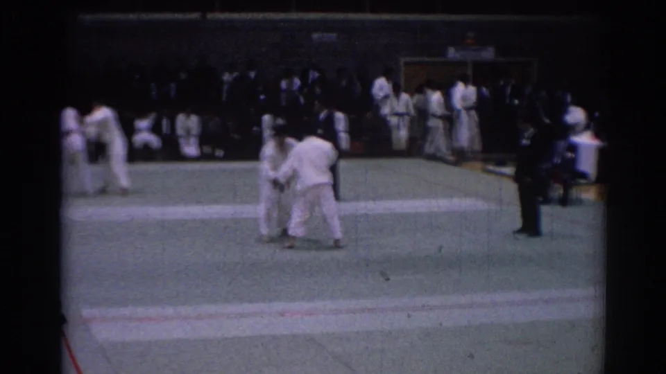 Indoor 1967 Los Angeles karate match frame with competitors in motion, a nearby official, and spectators along the gym edge.