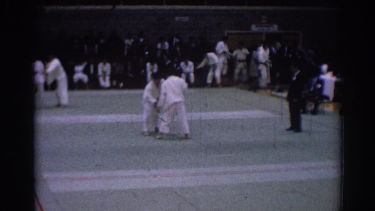 1967 Los Angeles home-movie frame showing two karate competitors on a green mat with seated spectators behind them.