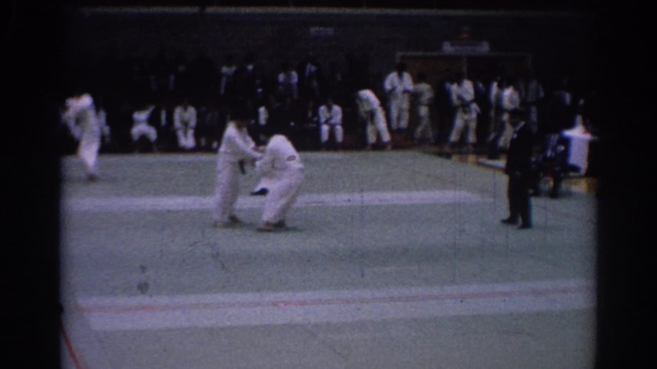 One karate competitor bends low while another stands nearby on the mat, with spectators in the background.