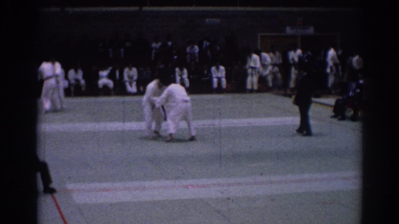 1967 Los Angeles indoor karate bout with two competitors grappling on a green mat and an official nearby.