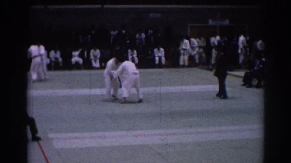 1967 Los Angeles indoor karate bout with two competitors grappling on a green mat and an official nearby.