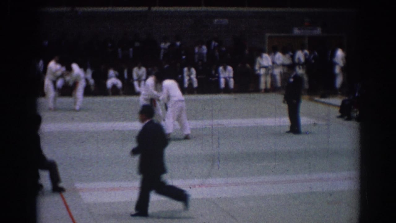 Man in a dark suit crosses the foreground during a 1967 Los Angeles karate match filmed on 8mm.