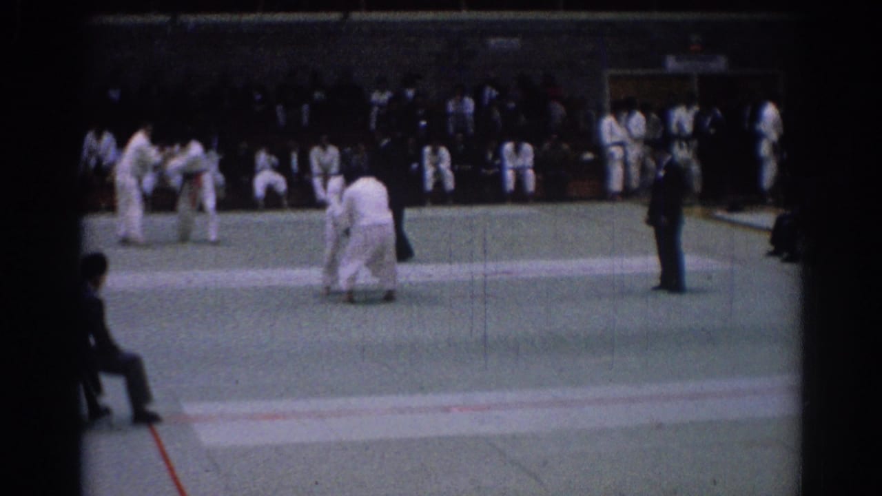 Two judo competitors spar on a gym mat while a referee watches nearby.