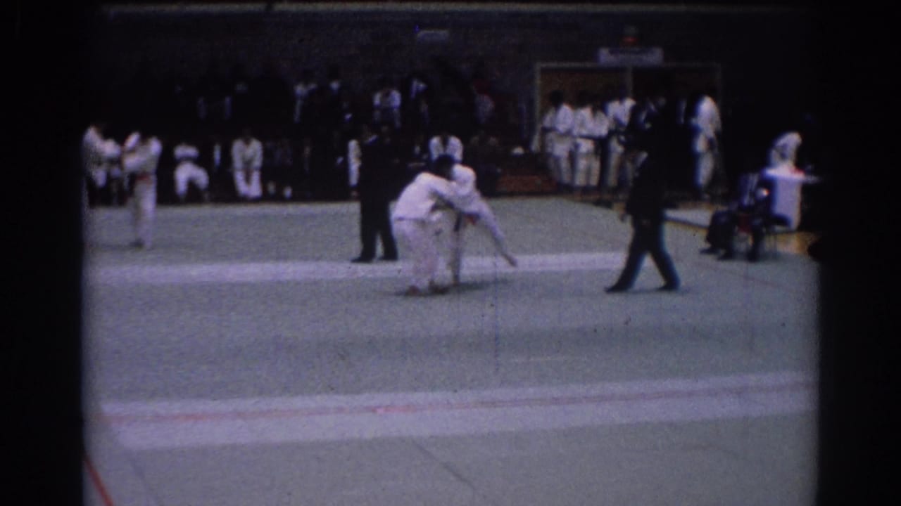 1967 Los Angeles judo tournament frame with competitors grappling on the mat, a referee in dark clothing, and uniformed participants along the wall.