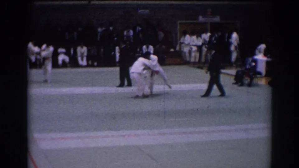 1967 Los Angeles judo tournament frame with competitors grappling on the mat, a referee in dark clothing, and uniformed participants along the wall.