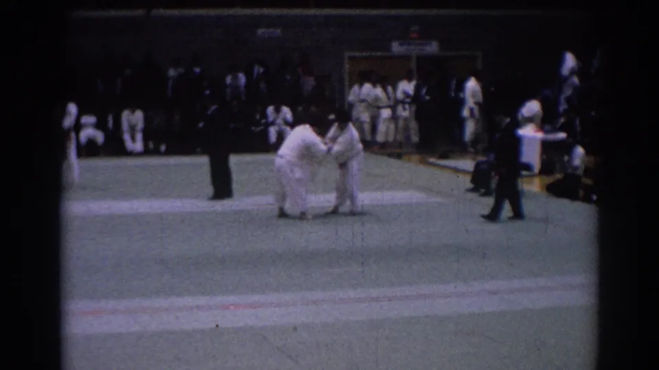 1967 Los Angeles judo tournament frame showing two white-gi competitors facing off, with a referee and several participants visible around the mat.